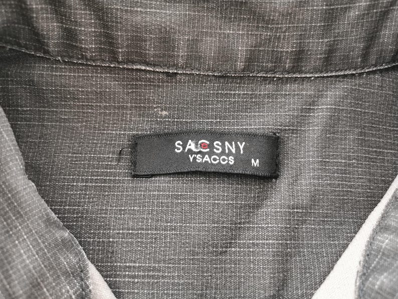 May include: Close-up of a gray collared shirt with a textured pattern. A black label with the text "SACSNY Y'SACCS M" is sewn on the shirt. The shirt is made of a woven fabric.
