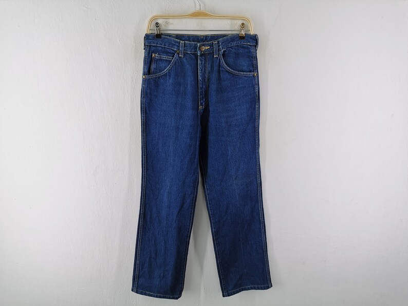 May include: A pair of classic blue denim jeans hanging on a wooden hanger. The jeans have a straight leg cut, five pockets, and a button and zipper closure. The denim is a medium wash, and the jeans appear to be new.