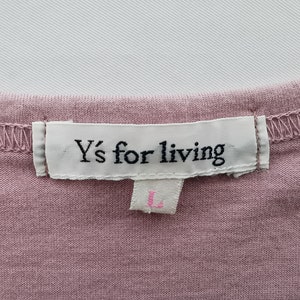 May include: Close-up of a dusty rose-colored garment with a white label stitched to the neckline. The label reads "Y's for living" in black text. Below the label is a smaller tag with the letter "L" in pink.