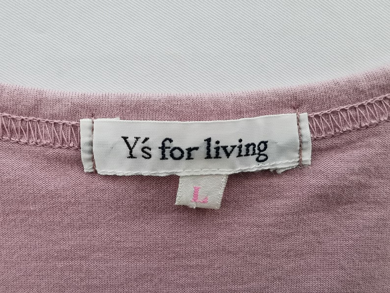 May include: Close-up of a dusty rose-colored garment with a white label stitched to the neckline. The label reads "Y's for living" in black text. Below the label is a smaller tag with the letter "L" in pink.