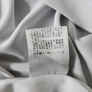 May include: Close-up of a white fabric with a tag attached. The tag has Japanese text. The fabric appears to be a light, breathable material, possibly for athletic wear or a similar garment.