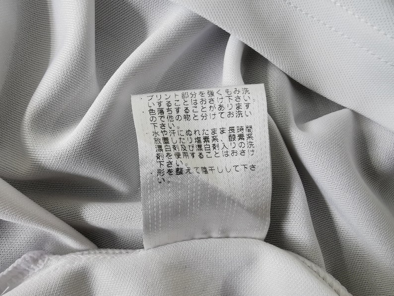 May include: Close-up of a white fabric with a tag attached. The tag has Japanese text. The fabric appears to be a light, breathable material, possibly for athletic wear or a similar garment.