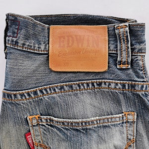 Puede incluir: Primer plano de vaqueros de mezclilla azul con costuras naranjas. Un parche de cuero marr&oacute;n en la cintura dice "EDWIN Exclusive Vintage". Los vaqueros tienen un dise&ntilde;o cl&aacute;sico de cinco bolsillos y una etiqueta roja Edwin.