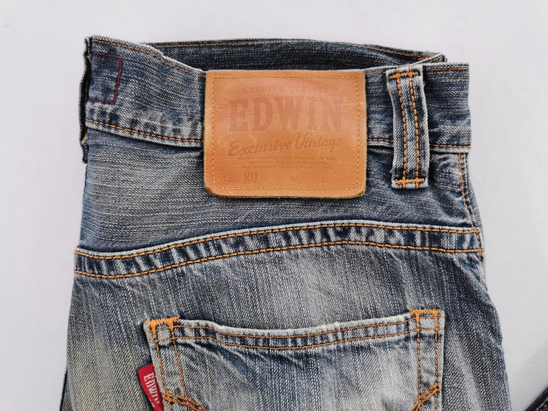 Puede incluir: Primer plano de vaqueros de mezclilla azul con costuras naranjas. Un parche de cuero marr&oacute;n en la cintura dice "EDWIN Exclusive Vintage". Los vaqueros tienen un dise&ntilde;o cl&aacute;sico de cinco bolsillos y una etiqueta roja Edwin.