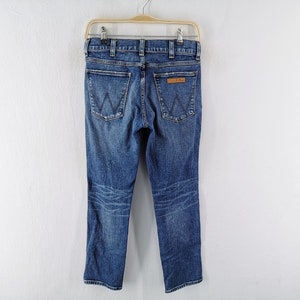 May include: Blue denim jeans with a classic five-pocket design, featuring the iconic "W" stitching on the back pockets and a leather patch. The jeans are hanging on a hanger against a white background.