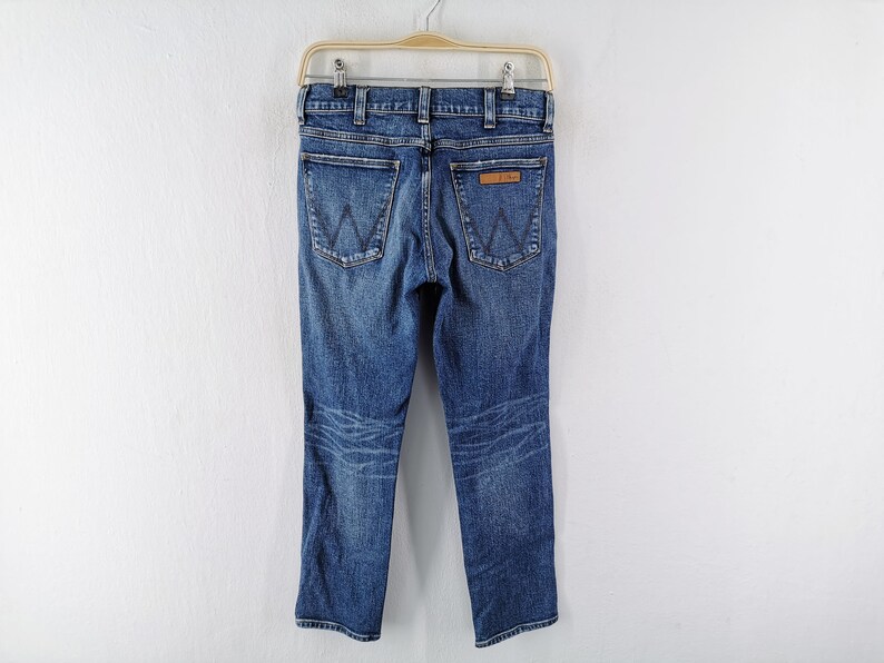 May include: Blue denim jeans with a classic five-pocket design, featuring the iconic "W" stitching on the back pockets and a leather patch. The jeans are hanging on a hanger against a white background.