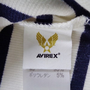 May include: A white and navy blue striped ribbed fabric garment featuring a white interior tag. The tag displays a gold winged star emblem above the text AVIREX. Below this, a secondary tag shows partial text and the percentage 5%.