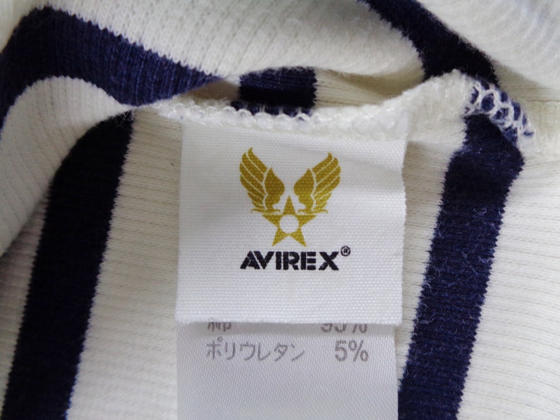 May include: A white and navy blue striped ribbed fabric garment featuring a white interior tag. The tag displays a gold winged star emblem above the text AVIREX. Below this, a secondary tag shows partial text and the percentage 5%.