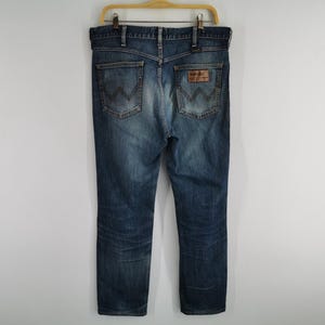 May include: Blue denim jeans with a classic five-pocket design, hanging on a wooden hanger. The jeans feature a faded wash and a leather patch on the back pocket. The back pockets have a distinctive 'W' stitching.