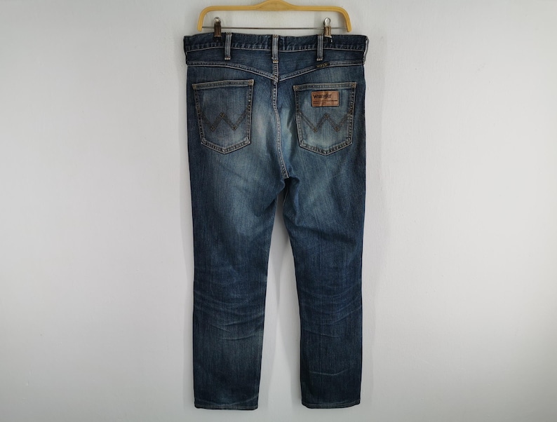 May include: Blue denim jeans with a classic five-pocket design, hanging on a wooden hanger. The jeans feature a faded wash and a leather patch on the back pocket. The back pockets have a distinctive 'W' stitching.