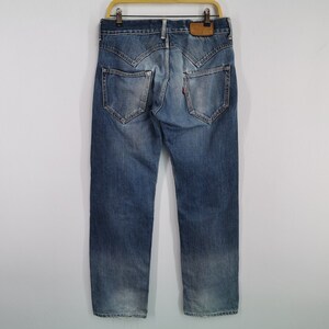 May include: Blue denim jeans with distinctive pocket and yoke designs. The jeans are viewed from the back, highlighting the stitching and fit. The jeans are suspended on a wooden hanger.