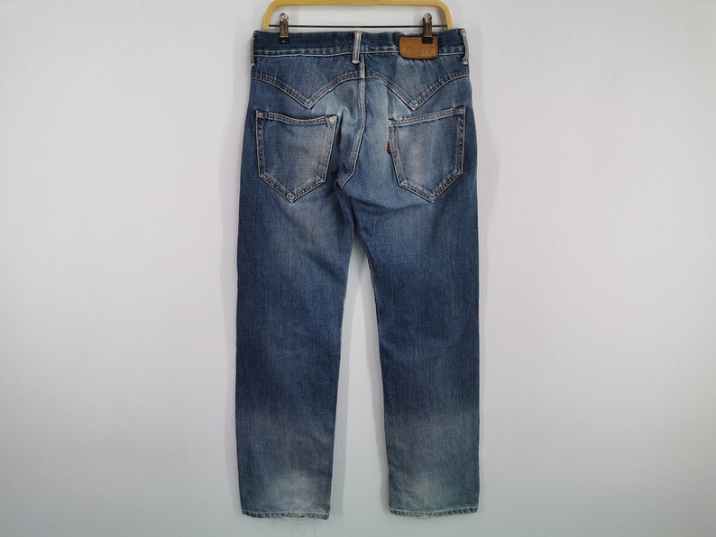 May include: Blue denim jeans with distinctive pocket and yoke designs. The jeans are viewed from the back, highlighting the stitching and fit. The jeans are suspended on a wooden hanger.