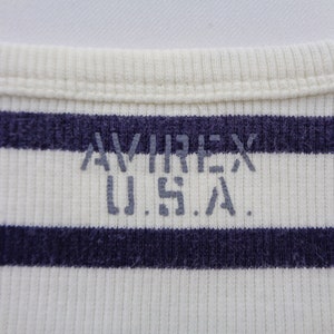 May include: A ribbed knit garment featuring horizontal navy blue and off-white stripes. The text AVIREX U.S.A. is printed in a distressed grey font across the center of the fabric, positioned below a ribbed crew neckline collar.