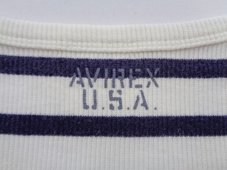 May include: A ribbed knit garment featuring horizontal navy blue and off-white stripes. The text AVIREX U.S.A. is printed in a distressed grey font across the center of the fabric, positioned below a ribbed crew neckline collar.