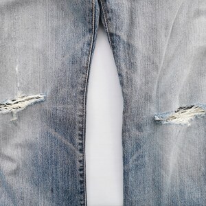 May include: Close-up of light blue denim jeans with distressed details. The jeans feature ripped areas at the knees, showcasing the frayed fabric underneath. The denim has a faded appearance, with lighter shades in the center.