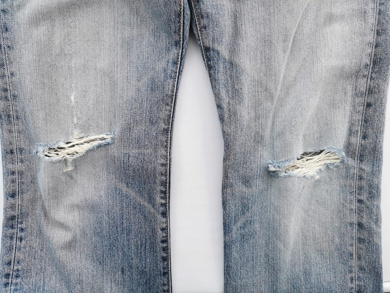 May include: Close-up of light blue denim jeans with distressed details. The jeans feature ripped areas at the knees, showcasing the frayed fabric underneath. The denim has a faded appearance, with lighter shades in the center.