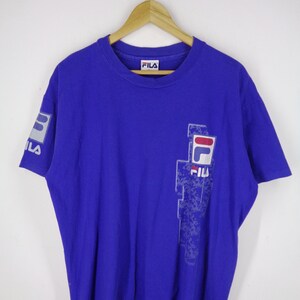 May include: A royal blue Fila t-shirt with a crew neck and short sleeves. The shirt features the Fila logo on the left sleeve and a larger design on the front, incorporating the brand's name and logo in white, red, and blue.