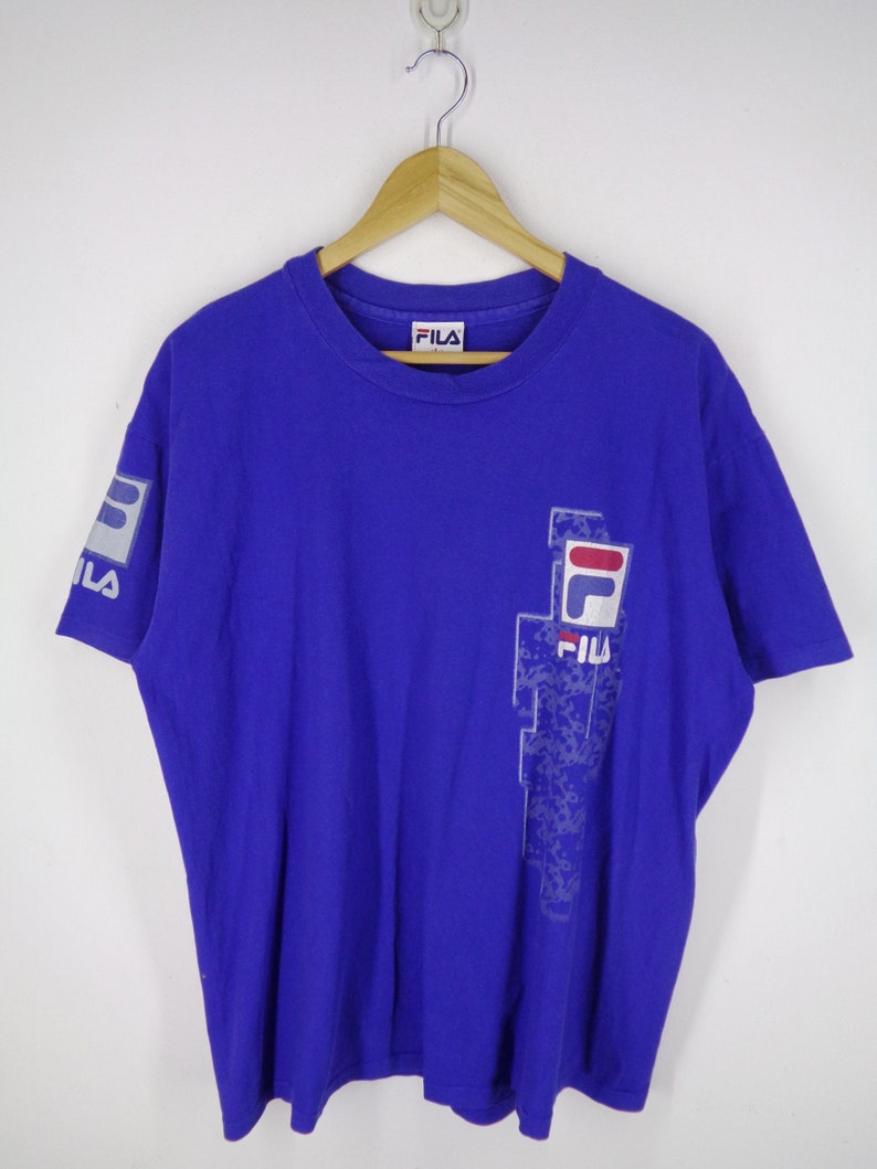 May include: A royal blue Fila t-shirt with a crew neck and short sleeves. The shirt features the Fila logo on the left sleeve and a larger design on the front, incorporating the brand's name and logo in white, red, and blue.