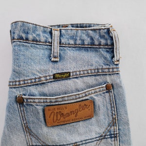 May include: Light blue denim Wrangler jeans with a classic five-pocket design. The jeans feature a brown leather patch on the back pocket with the Wrangler logo and the text "Blue Bell's Authentic Western Jeans".