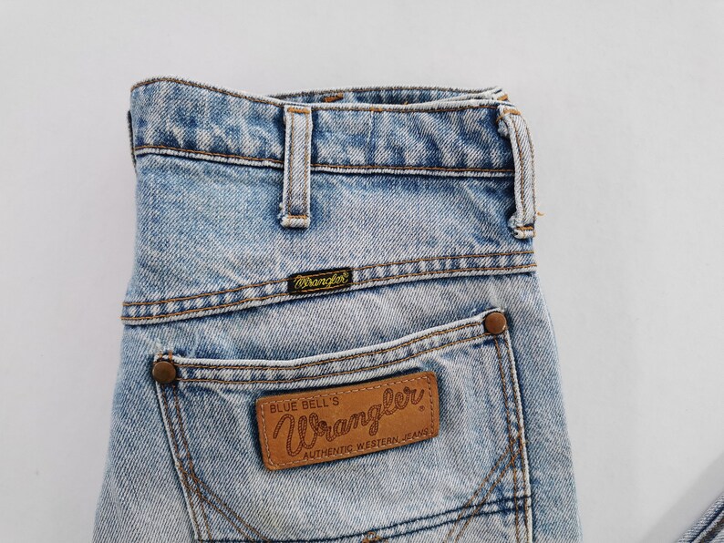 May include: Light blue denim Wrangler jeans with a classic five-pocket design. The jeans feature a brown leather patch on the back pocket with the Wrangler logo and the text "Blue Bell's Authentic Western Jeans".