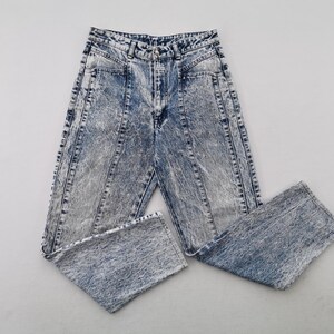 May include: Acid wash denim jeans with a high waist and straight leg. The jeans have a light blue and gray marbled effect, with vertical seams down the front and back of the legs. The jeans are folded.