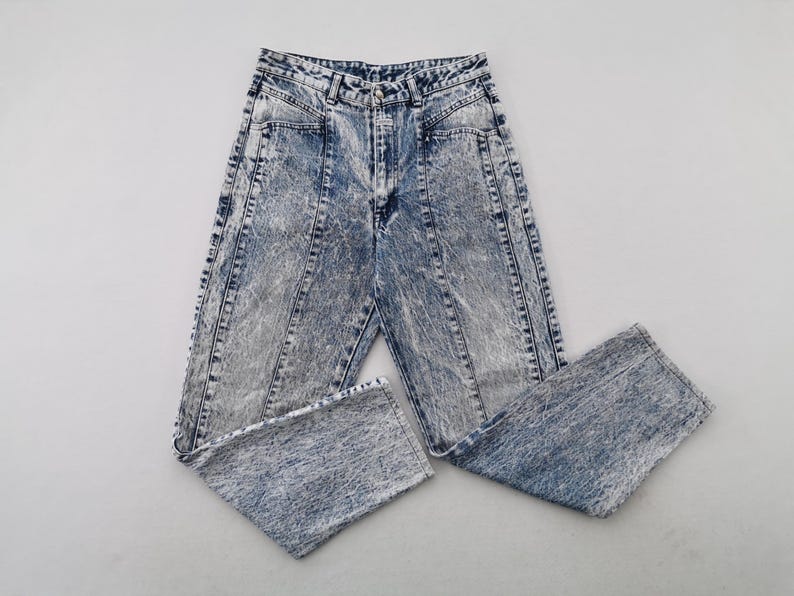 May include: Acid wash denim jeans with a high waist and straight leg. The jeans have a light blue and gray marbled effect, with vertical seams down the front and back of the legs. The jeans are folded.