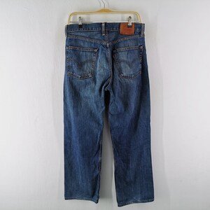 May include: Blue denim jeans hanging on a wooden hanger. The jeans have a classic five-pocket design, with a leather patch on the back. The denim has a faded, worn look.