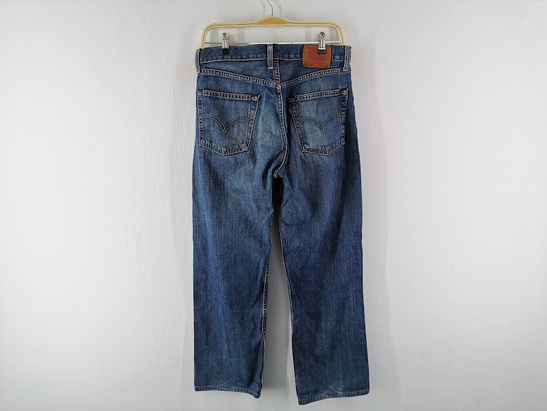 May include: Blue denim jeans hanging on a wooden hanger. The jeans have a classic five-pocket design, with a leather patch on the back. The denim has a faded, worn look.