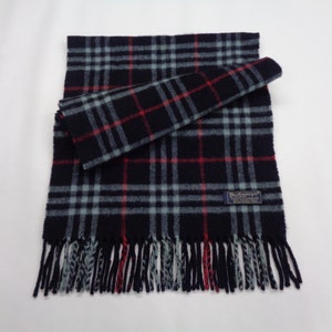 May include: A navy blue scarf with a plaid pattern in light blue and red. The scarf has a fringed edge and a small label with text. The scarf is folded and displayed on a white background.