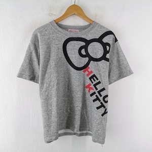 May include: A heather grey short-sleeve t-shirt featuring a large black outline of a bow graphic on the upper right side. The text HELLO KITTY is printed vertically in black and red letters below the bow. The shirt has a crew neckline.