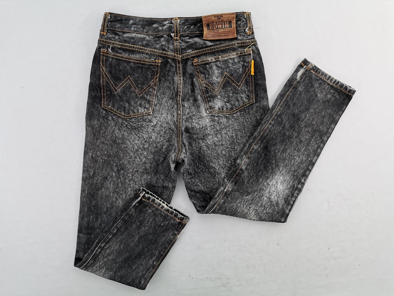 May include: Acid wash black denim jeans with orange stitching and a classic five-pocket design. The jeans feature the Edwin brand label on the back and a yellow tag. A stylish and timeless addition to any wardrobe.