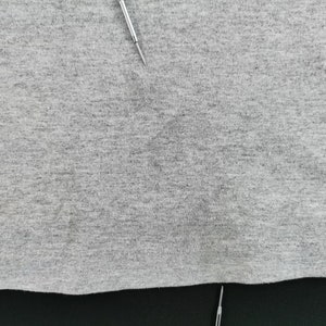 May include: Close-up of a gray fabric with a textured surface. Two silver pins are inserted into the fabric. The fabric appears to be a light gray color.