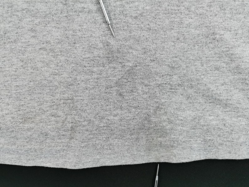 May include: Close-up of a gray fabric with a textured surface. Two silver pins are inserted into the fabric. The fabric appears to be a light gray color.