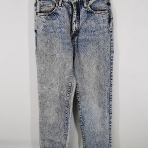 K&ouml;nnte beinhalten: S&auml;uregewaschene Jeans aus Denim, die an einem goldenen Kleiderb&uuml;gel h&auml;ngen. Die Jeans haben eine hohe Taille, einen Rei&szlig;verschluss und ein klassisches F&uuml;nf-Taschen-Design. Der Denim ist eine Mischung aus Blau und Grau mit einem verblichenen, strukturierten Aussehen.