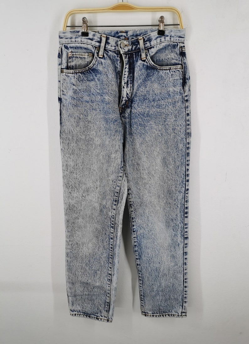 K&ouml;nnte beinhalten: S&auml;uregewaschene Jeans aus Denim, die an einem goldenen Kleiderb&uuml;gel h&auml;ngen. Die Jeans haben eine hohe Taille, einen Rei&szlig;verschluss und ein klassisches F&uuml;nf-Taschen-Design. Der Denim ist eine Mischung aus Blau und Grau mit einem verblichenen, strukturierten Aussehen.