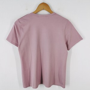 May include: A short-sleeved, mauve-colored t-shirt hanging on a wooden hanger. The shirt is made of a soft material and has a simple, classic design. The background is a plain white wall.