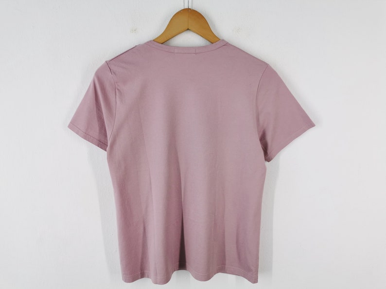May include: A short-sleeved, mauve-colored t-shirt hanging on a wooden hanger. The shirt is made of a soft material and has a simple, classic design. The background is a plain white wall.