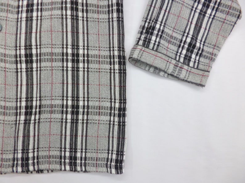 May include: Close-up of a gray flannel shirt with a classic plaid pattern. The pattern features black, white, and thin red lines. The sleeve cuff is visible, showing the same plaid design. The shirt is buttoned and appears to be made of a soft fabric.