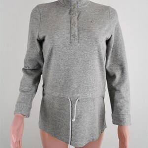 May include: A gray long-sleeve shirt with a high collar and a button-down placket. The shirt has a drawstring at the waist and a small logo on the chest. The sleeves have ribbed cuffs.