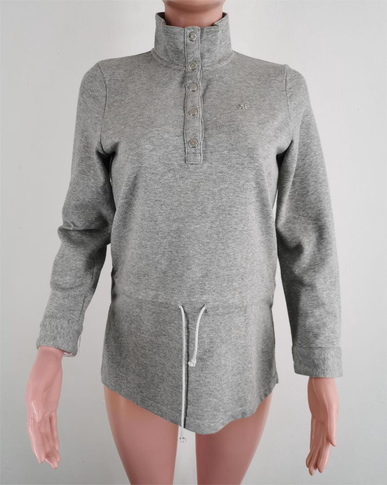 May include: A gray long-sleeve shirt with a high collar and a button-down placket. The shirt has a drawstring at the waist and a small logo on the chest. The sleeves have ribbed cuffs.