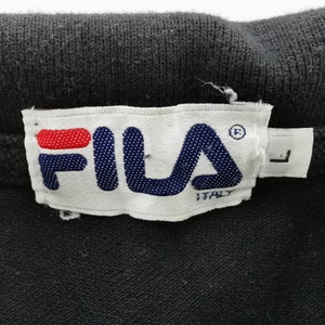 May include: Close-up of a black garment with a white tag featuring the FILA logo in blue and red. The tag also includes the word "ITALY" and the size "L".