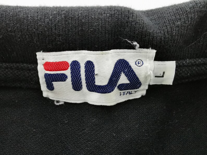 May include: Close-up of a black garment with a white tag featuring the FILA logo in blue and red. The tag also includes the word "ITALY" and the size "L".