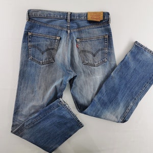 Puede incluir: Vaqueros de mezclilla azul con un lavado desgastado, dos bolsillos traseros con costuras arqueadas, una etiqueta de cuero color canela en la cintura y una pequeña etiqueta roja en el bolsillo derecho. El pantalón tiene un diseño clásico.