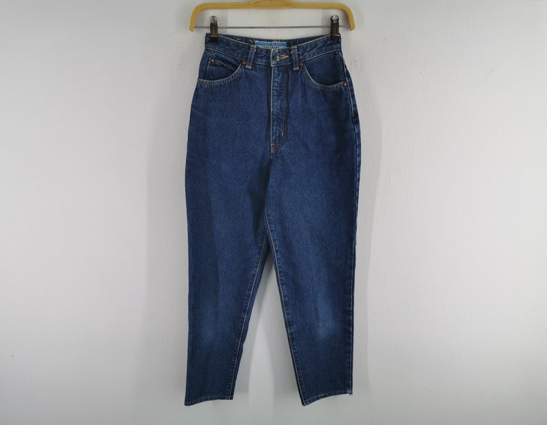 May include: Dark blue denim jeans hanging on a wooden hanger. The jeans have a high waist, pockets, and a button and zipper closure. The denim appears to be a medium weight, with a classic, straight-leg fit.