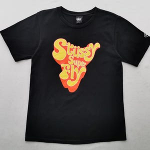 May include: Black t-shirt featuring the text "Stussy Super Fly" in a yellow and orange retro design. The short-sleeved top has a crew neck and a small logo on the upper left chest. A graphic is on the left sleeve.