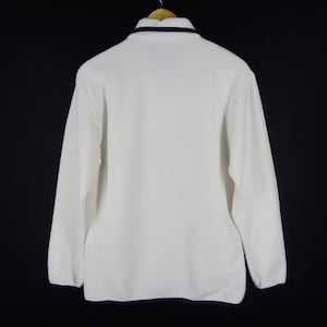 May include: White long-sleeved fleece pullover with a navy blue trim around the collar. The back view of the shirt is displayed against a black background. The shirt is hanging on a wooden hanger.
