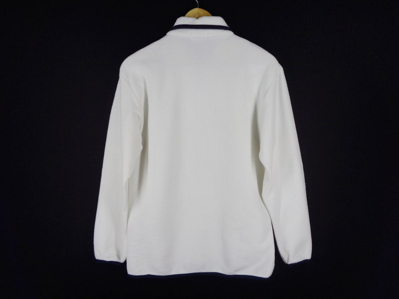 May include: White long-sleeved fleece pullover with a navy blue trim around the collar. The back view of the shirt is displayed against a black background. The shirt is hanging on a wooden hanger.