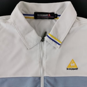 May include: White golf jacket with a full zipper and a collared neckline. The jacket features blue and yellow accents on the collar and a blue striped pattern at the bottom. The Le Coq Sportif logo is visible on the chest.