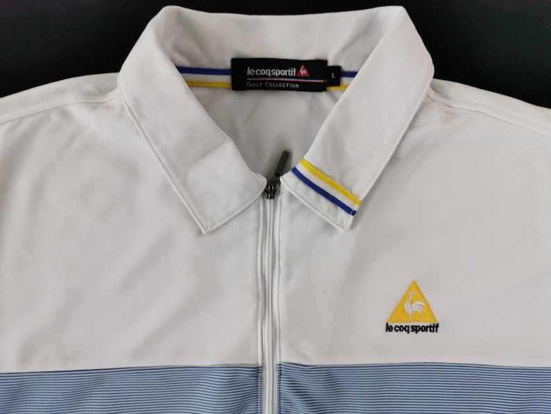 May include: White golf jacket with a full zipper and a collared neckline. The jacket features blue and yellow accents on the collar and a blue striped pattern at the bottom. The Le Coq Sportif logo is visible on the chest.
