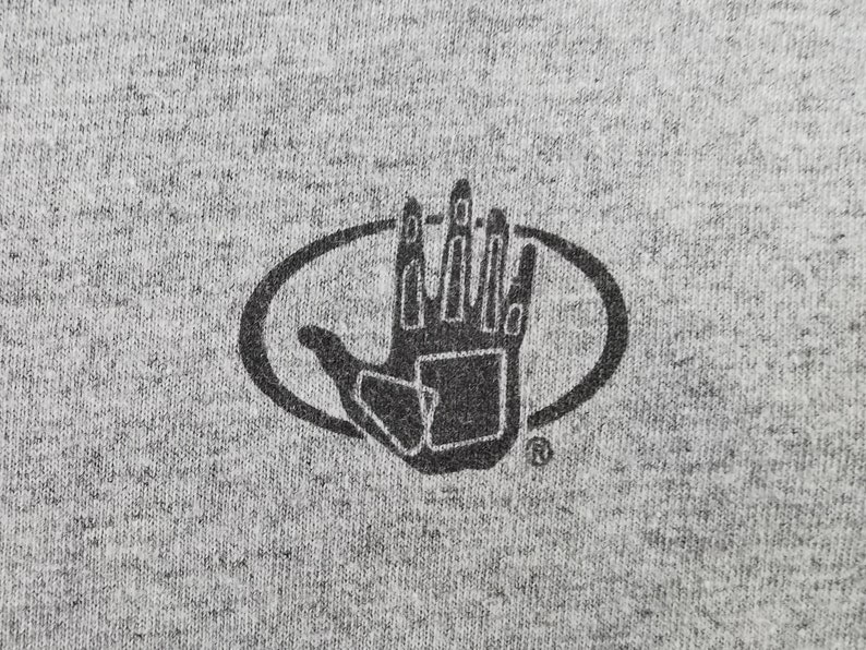 May include: Close-up of a gray t-shirt with a black graphic. The graphic is a hand inside an oval, with the fingers and palm detailed. The t-shirt has a heathered texture.